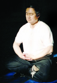 a photograph of Master Shouyu Liang meditating. He sits in a half lotus position, illuminated by sunlight, framed by natural shadow.